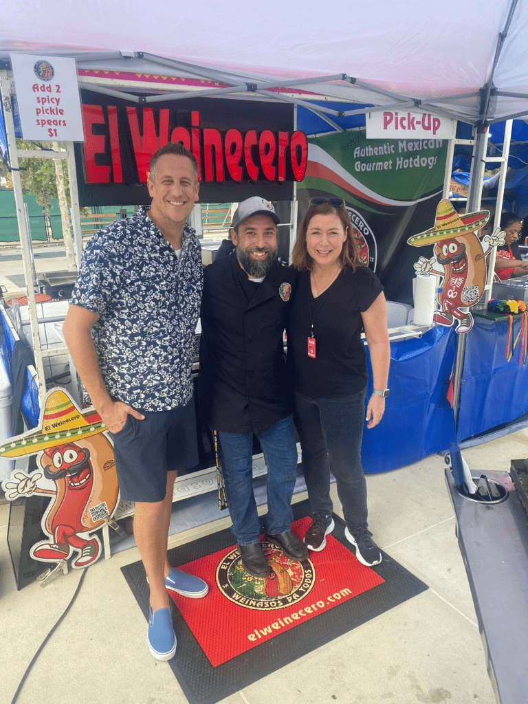 Three people smiling at a food stall.