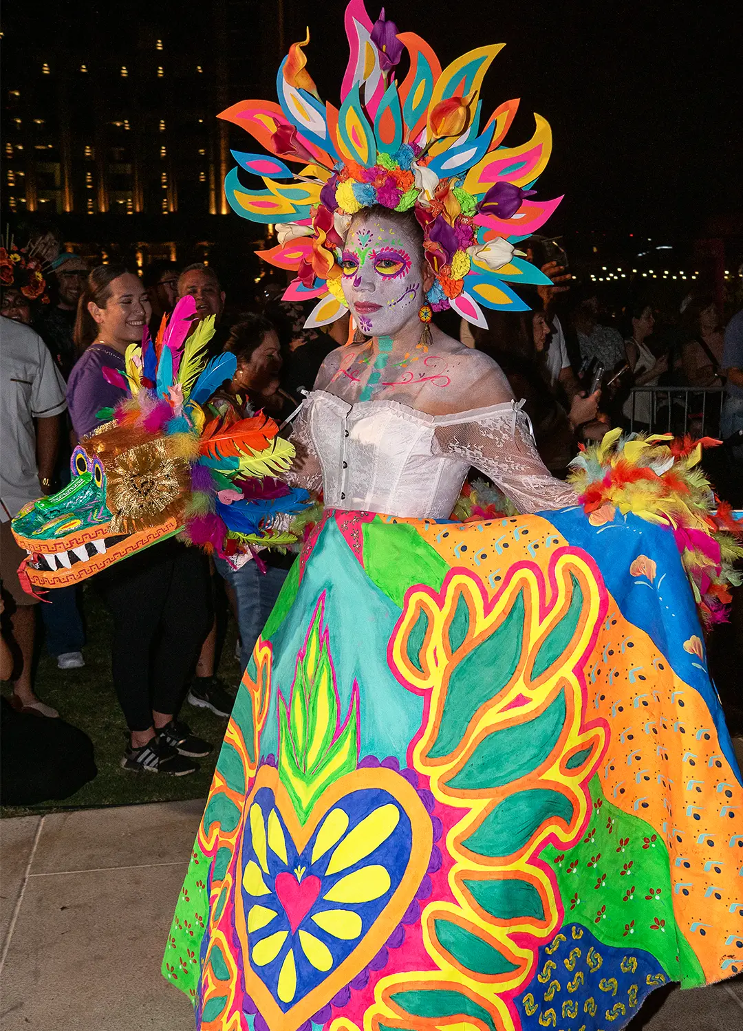 Catrina procession participant in bright neon colors with a large headpiece and hand-painted skirt among crowds at night at Muertos Fest