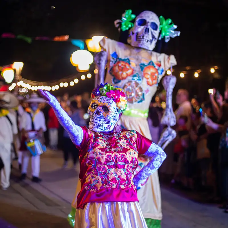 Colorfully dressed procession participant with calavera makeup and illuminated puppet among crowds at night during Muertos Fest