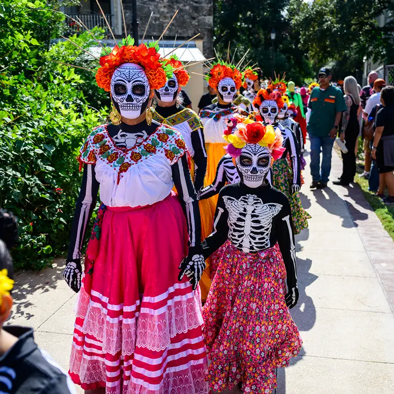 Adult and child Catrinas in colorful skirts and flower crowns walking together in the Muertos Fest procession