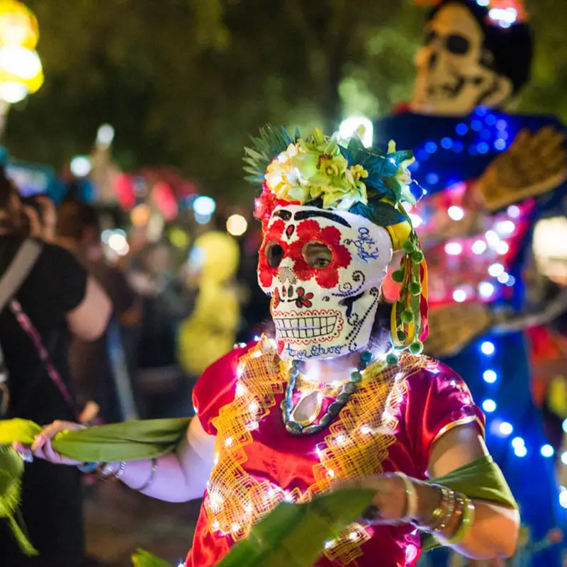 Young procession participant in colorful calavera attire illuminated at night among crowds at Muertos Fest