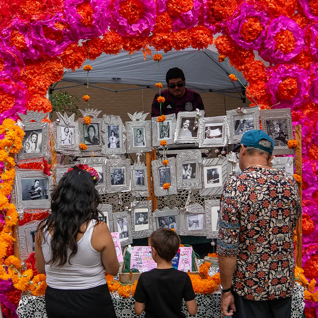 Family viewing a Día de los Muertos ofrenda filled with photographs of loved ones and large pink and orange flower