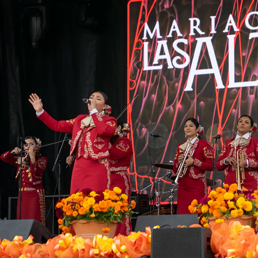 Mariachi Las Alteñas, an all-women mariachi group, performing on stage surrounded by marigolds at Muertos Fest