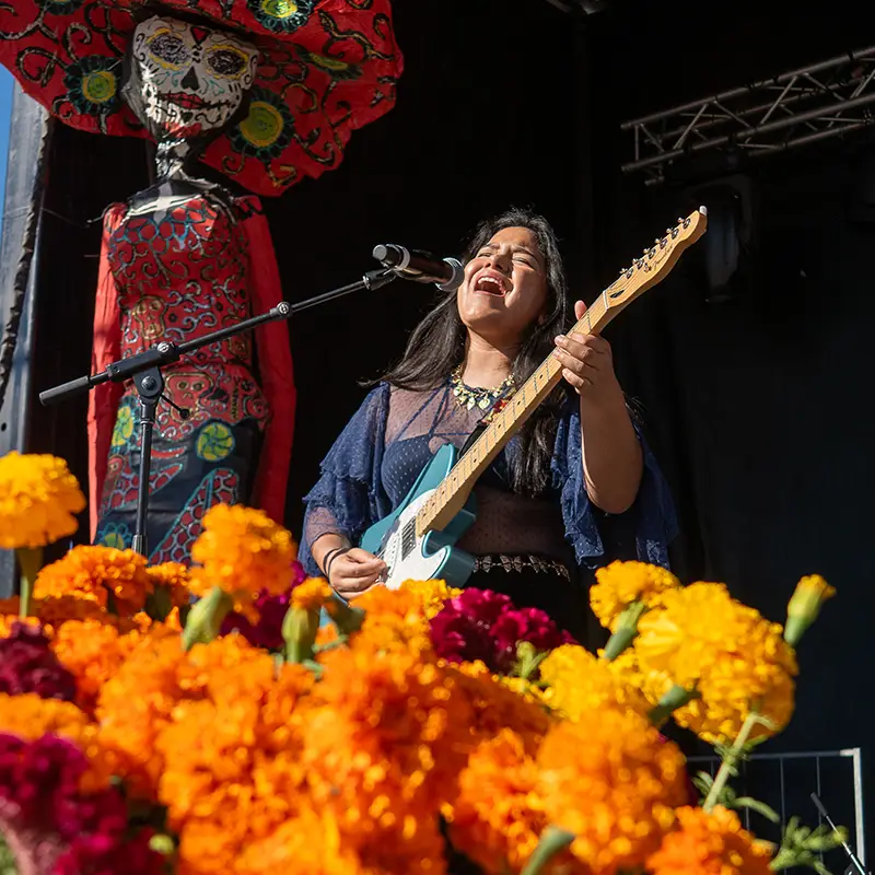 Musician performing among marigolds and Catrina imagery during the Día de los Muertos celebration at Muertos Fest on main stage