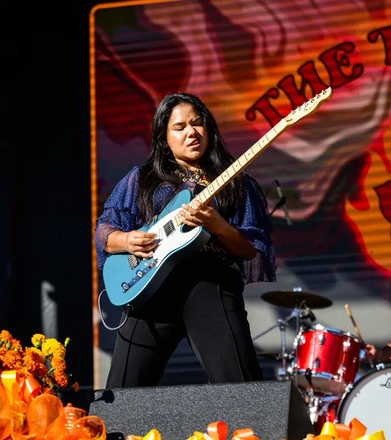Female guitarist performing live on stage surrounded by marigolds at Muertos Fest